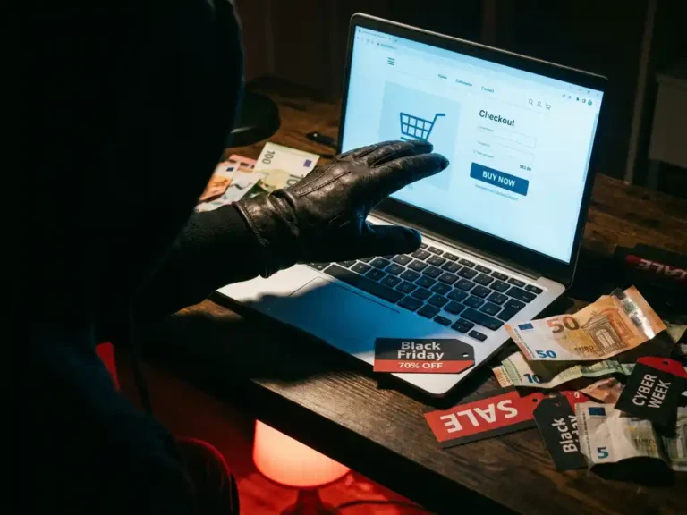 Behandschuhte Hand greift zu leuchtendem Laptop mit Checkout-Seite, umgeben von Black-Friday-Tags und Euroscheinen auf dunklem Schreibtisch.