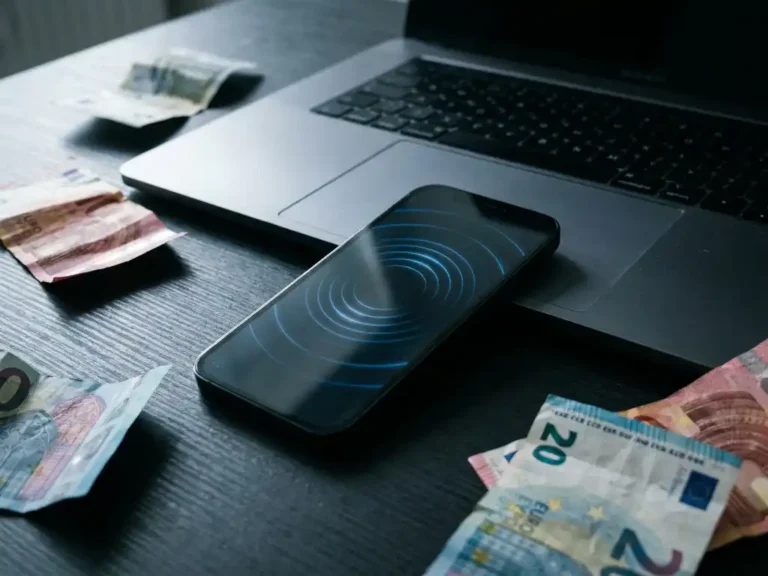 Smartphone und Laptop auf dunklem Schreibtisch mit visualisierten Ghost-Click-Wellen auf dem Bildschirm, umgeben von zerknüllten Euroscheinen.