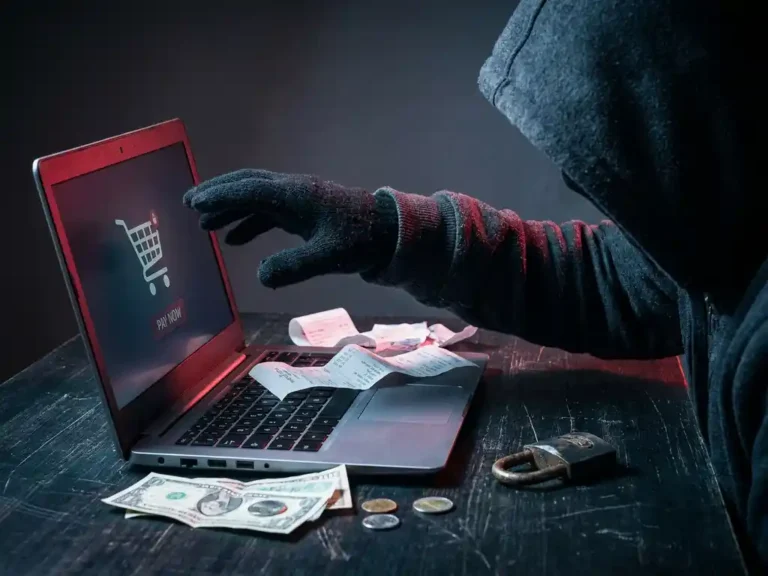 Vermummte Person mit Hand über leuchtendem Laptop mit Online-Warenkorb, zerbrochenes Vorhängeschloss auf dunklem Schreibtisch.