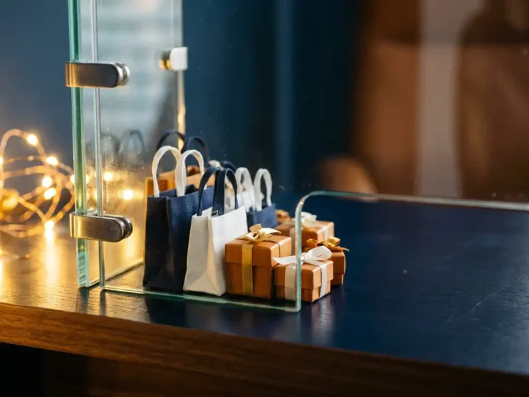 Glasschild schützt Miniatur-Einkaufstaschen und Geschenkboxen auf modernem Schreibtisch, goldenes Licht, Marineblau und Bernsteintöne.