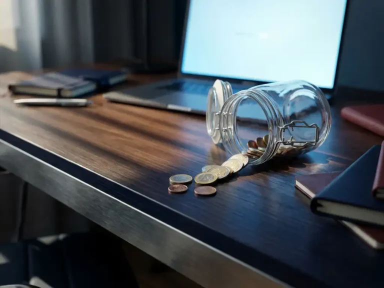 Leeres Glasgefäß auf der Seite liegend mit verstreuten Euromünzen auf modernem Schreibtisch, Laptop im Hintergrund.